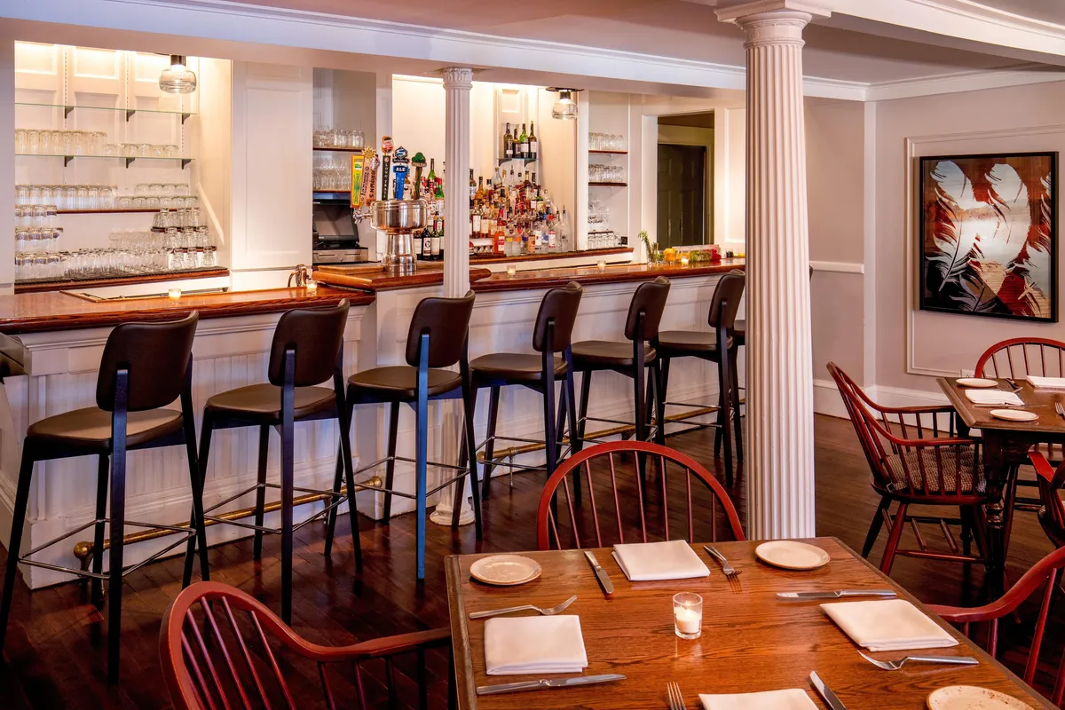 This interior image features a welcoming bar counter with comfortable stools, elegant table settings, and local artwork adorning the walls. Guests are seen socializing, dining, and enjoying drinks in a relaxed, inviting atmosphere.
