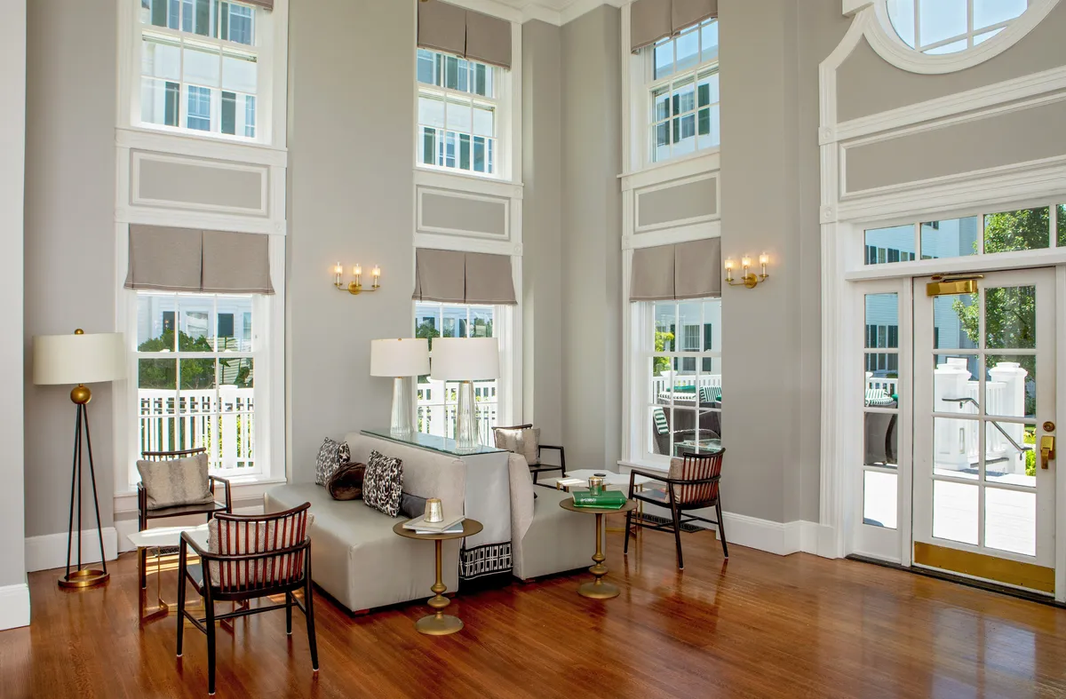 This image showcases a sophisticated and inviting interior space with large windows that fill the room with natural light. A comfortable seating arrangement, decorative mirror, and elegant light fixtures set a refined yet cozy tone. The space feels welcoming and versatile, ideal for reading, relaxing, or socializing.