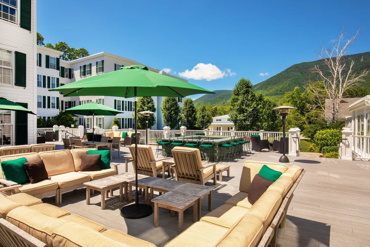 An exterior dining area featuring green umbrellas and plush seating, set against a picturesque mountain backdrop. Guests relax and socialize while savoring meals outdoors, surrounded by Vermont's natural beauty.