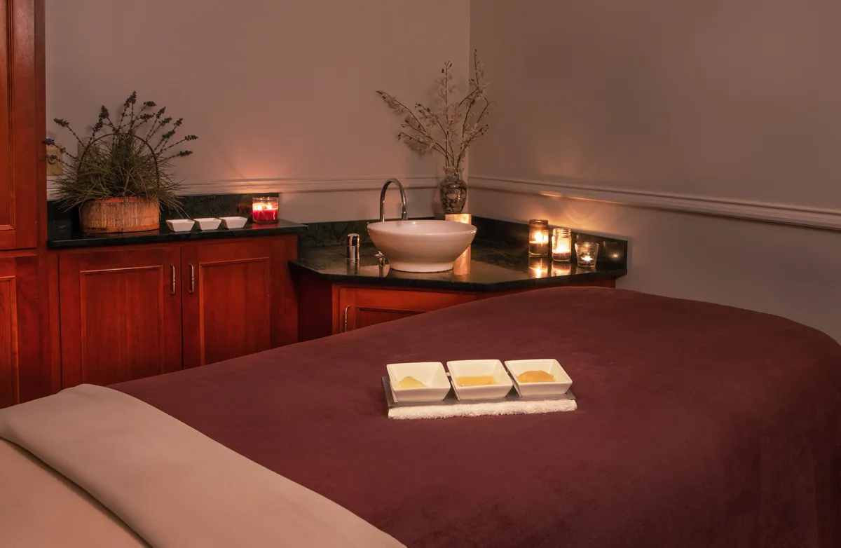 The image displays a serene spa-like interior featuring a massage table, a bowl filled with oils, softly glowing candles, and a decorative plant. The lighting is gentle, and the space exudes peace and relaxation, reminiscent of a high-end wellness sanctuary.