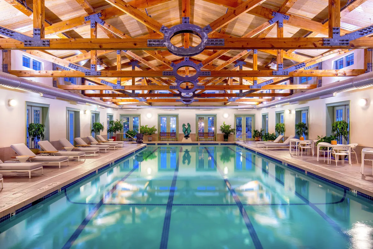 A luxurious indoor swimming pool lined with wooden beams, soft ambient lighting, and comfortable lounge chairs. Decorative plants add a touch of greenery, and the serene atmosphere suggests both relaxation and opportunities for wellness activities. The pool area feels inviting and upscale, suitable for socializing, exercising, or unwinding after a day of adventure.