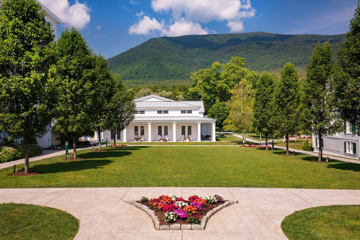 An inviting exterior shot of a manicured lawn with a vibrant flower bed, a stately white building, and a stunning mountain backdrop. The scene captures opportunities for outdoor activities and recreation, such as lawn games and social gatherings, while emphasizing the natural beauty that surrounds the resort.