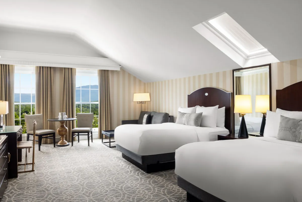 The image features a bright, elegant interior with two plush beds, a stylish desk and chairs, large sunlit windows offering scenic views, a decorative lamp, and a mirrored wall, all arranged to maximize comfort and sophistication. The space is inviting, spacious, and ideal for relaxation or productivity, with New England charm and resort luxury evident in every detail.